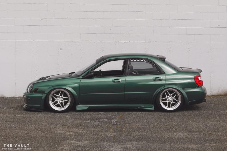 May include: A dark green sedan with custom silver alloy wheels and a lowered suspension. The car has a spoiler and tinted windows, parked in front of a white brick wall. The car's body is a deep emerald green.