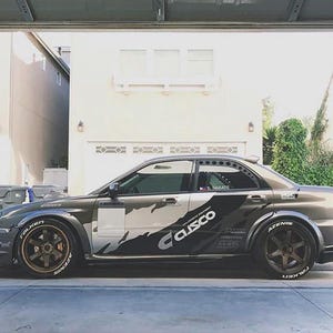 May include: A gray and silver sports car with custom decals and wide tires. The car features the "Cusco" logo and "Falken Azenis" tire branding. The vehicle is parked in a driveway.