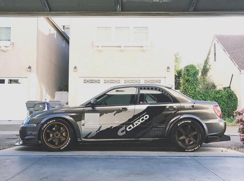 May include: A gray and silver sports car with custom decals and wide tires. The car features the "Cusco" logo and "Falken Azenis" tire branding. The vehicle is parked in a driveway.