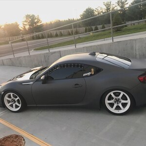 May include: A dark grey sports car with white wheels is parked on a concrete surface. The car has a spoiler and a sticker on the rear window. The car is angled towards the left, with the sun setting in the background.