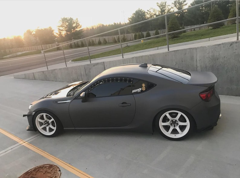 May include: A dark grey sports car with white wheels is parked on a concrete surface. The car has a spoiler and a sticker on the rear window. The car is angled towards the left, with the sun setting in the background.