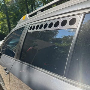 May include: A grey SUV with a black metal window guard featuring multiple circular cutouts. The vehicle has a roof rack and tinted windows, parked outdoors on a sunny day. The guard is mounted above the rear passenger window.
