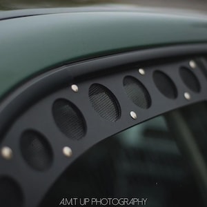 May include: Close-up of a dark green car with a black window visor. The visor has multiple circular openings with mesh inserts and is secured with silver rivets. The car's roof is visible.