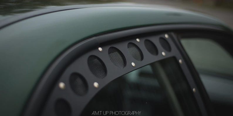 May include: Close-up of a dark green car with a black window visor. The visor has multiple circular openings with mesh inserts and is secured with silver rivets. The car's roof is visible.