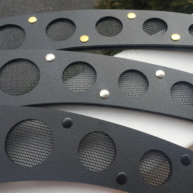May include: Close-up of three black metal decorative pieces. Each piece has circular mesh inserts and is adorned with silver and gold-colored rivets. The pieces have a curved shape and a textured surface.