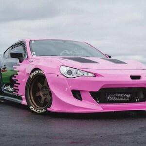 May include: A modified pink sports car with black detailing and a body kit. The car has custom wheels, a front splitter, and the word "Vortech" on the front. The car has the word "Certified" on the side.