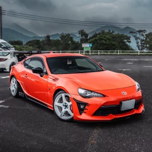 May include: Vibrant orange sports car with white alloy wheels and black detailing. The car features a rear spoiler and is parked on tarmac. The Toyota badge is visible.