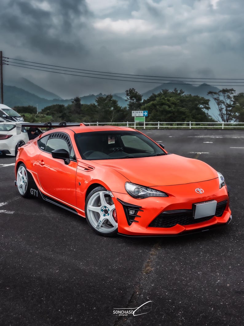 May include: Vibrant orange sports car with white alloy wheels and black detailing. The car features a rear spoiler and is parked on tarmac. The Toyota badge is visible.