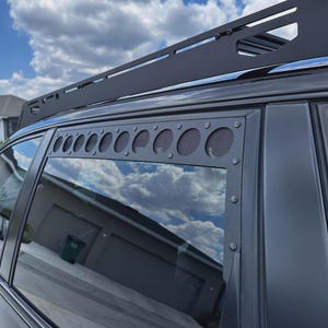 May include: Black vehicle roof rack and window vent covers. The rack is a flat, rectangular design with a series of crossbars. The window vent covers have a series of circular openings with mesh backing. The sky and clouds are reflected in the window.