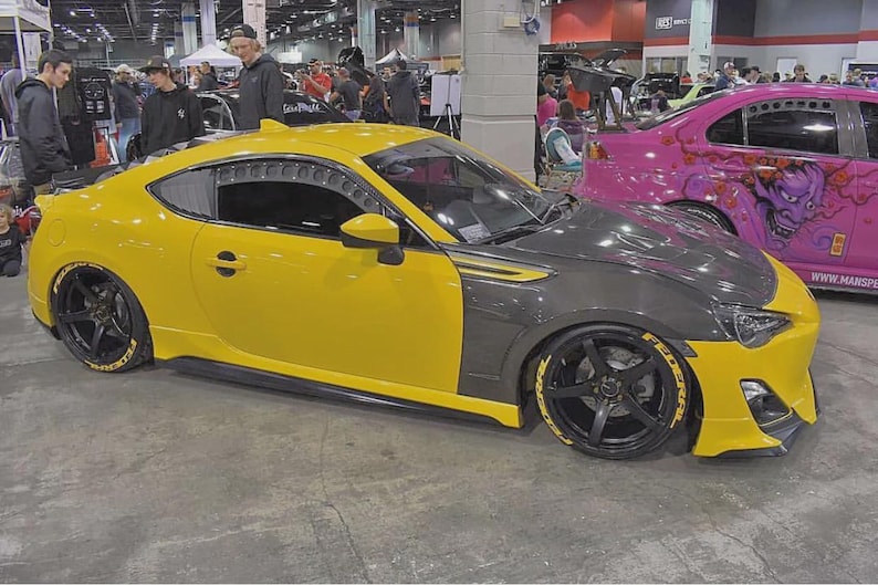 May include: A yellow sports car with a grey bonnet and black wheels. The car has a custom body kit and is parked in a convention centre. The wheels have yellow lettering.