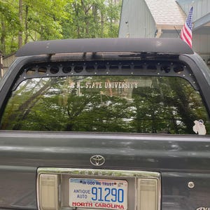 Peut inclure: Un Toyota 4Runner gris avec un porte-bagages noir et un autocollant sur la lunette arrière indiquant "N.C. STATE UNIVERSITY". Le véhicule a une plaque d'immatriculation de Caroline du Nord et un drapeau américain fixé sur le toit.