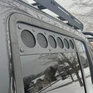 May include: Black metal vehicle window vent cover with multiple circular mesh openings. The cover is mounted on a black vehicle, with a roof rack visible above. The background shows a snowy outdoor scene.
