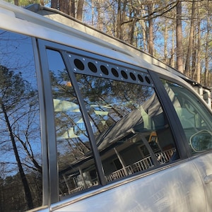 May include: A side view of a silver SUV with a black window visor featuring multiple circular cutouts. The windows reflect trees and a house. The vehicle is parked outside on a sunny day.
