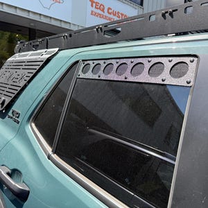 May include: A teal SUV with black accessories, including a roof rack and window vents. The window vent features a series of circular openings with mesh, and the rear window has a metal vent cover with the text "TEQ CUSTOM".