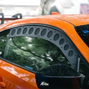 May include: Close-up of an orange sports car with a black window visor featuring multiple circular cutouts. The car's side mirror has the word "Mitsa" printed on it. The car is wet, suggesting it is raining.