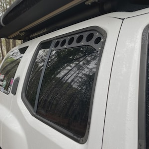 May include: A white SUV with a black window frame and a black roof rack. The rear passenger window has a custom black metal panel with circular cutouts. The background shows trees and a glimpse of a house.