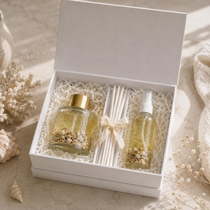 May include: A white gift box containing a glass diffuser bottle with a gold cap, a glass spray bottle with a white pump, and a bundle of white reed sticks tied with a beige ribbon. Both glass bottles contain a clear liquid and small beige stones.