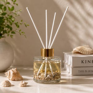 May include: A clear glass reed diffuser bottle with a gold-colored collar, filled with a light-colored liquid and small seashells at the base. Four white reed sticks are inserted into the bottle, extending upwards from the center opening.