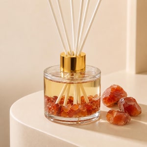 May include: A clear glass reed diffuser bottle with a gold-colored metal collar, filled with a light amber liquid and small, polished orange-red carnelian stones at the base. Several white reed sticks extend upward from the center of the bottle.