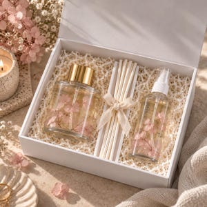 May include: A white gift box containing a clear glass diffuser bottle with a gold cap, a bundle of white reed sticks tied with a cream ribbon, and a clear glass spray bottle, all filled with liquid and pink hydrangea flower petals.