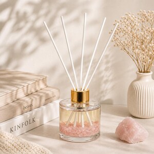 May include: A clear glass reed diffuser bottle with a gold collar, filled with light-coloured liquid and small pink crystals at the base. Five white reed sticks are inserted into the bottle, which is positioned near books and a pink stone.