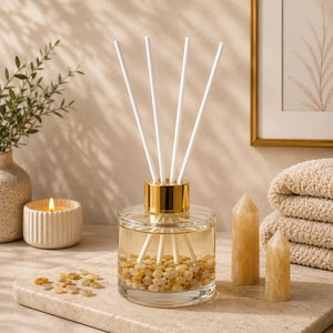 May include: A clear glass reed diffuser bottle with a gold-toned collar, filled with light-coloured liquid and small yellow stones at the base. Four white reed sticks are inserted into the bottle, extending upwards from the centre opening.