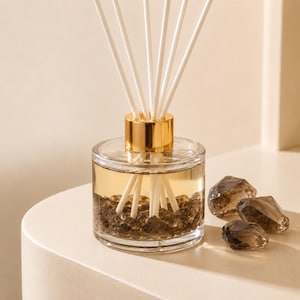 May include: A clear glass reed diffuser bottle with a gold-colored metal collar, filled with light amber liquid and dark brown decorative stones at the base. Six white reed sticks extend from the top, accompanied by three loose dark brown crystals.