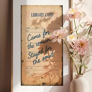 May include: Framed vintage-style print with the text "LIBRARY CARD" and "Came for the roman, Stayed for the smut." The print has a distressed, aged appearance. Pink flowers in a white vase are in the foreground.