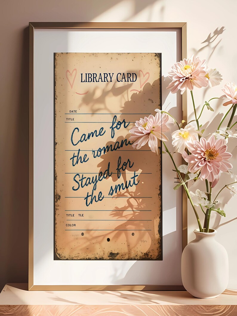 May include: Framed vintage-style print with the text "LIBRARY CARD" and "Came for the roman, Stayed for the smut." The print has a distressed, aged appearance. Pink flowers in a white vase are in the foreground.