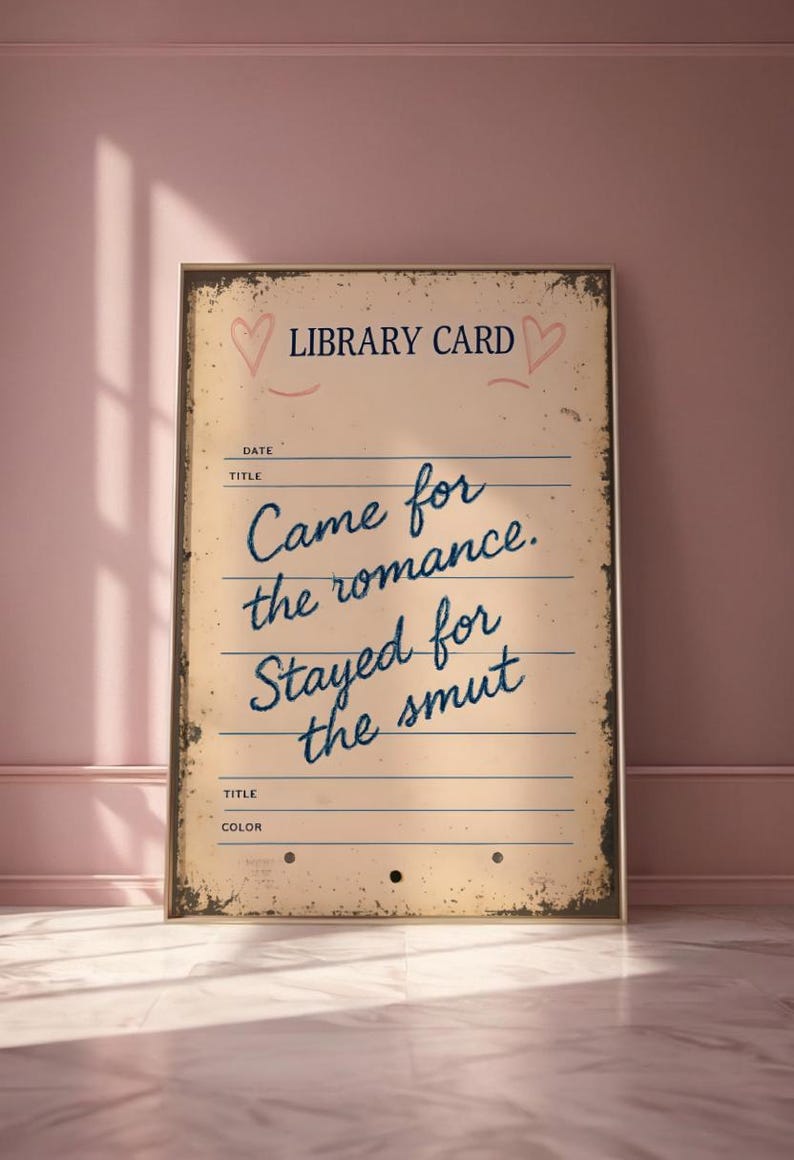 May include: A framed vintage-style library card print with the text "Came for the romance. Stayed for the smut" in blue script. The print has a distressed, aged appearance with hearts and lines.