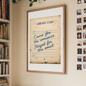 A framed wall art print that says ‘Came for the romance, stayed for the smut’, hanging on the wall above the bed, surrounded by plants and a bookcase
