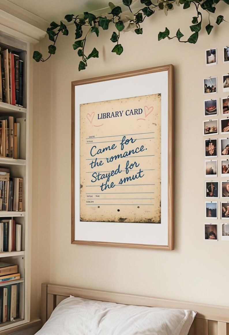 A framed wall art print that says ‘Came for the romance, stayed for the smut’, hanging on the wall above the bed, surrounded by plants and a bookcase