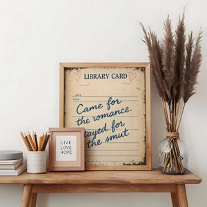 A framed wall art print that says ‘Came for the romance, stayed for the smut’. Styled on a side table with some pencils and plants