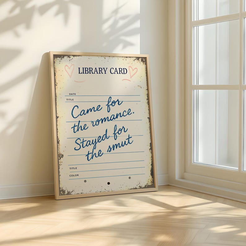 A framed wall art print that says ‘Came for the romance, stayed for the smut’. Styled in a light room near window with white walls and light wooden floors..