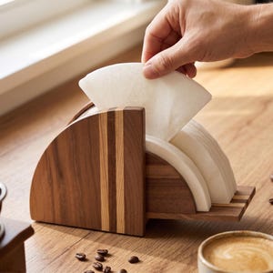 May include: A wooden coffee filter holder with a semi-circular design. The holder is made of dark brown wood with lighter wood accents. White paper coffee filters are stacked inside. A hand is reaching in to grab a filter. Coffee beans and a cup of coffee are in the background.