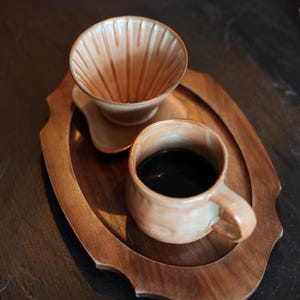 May include: A ceramic coffee pour-over set with a mug filled with dark coffee, resting on a wooden tray. The pour-over has a ridged interior. The mug and pour-over are a light brown color.