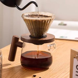 Könnte beinhalten: Ein Glas-Pour-Over-Kaffeebereiter mit Holzsockel und Griff. Heißes Wasser wird über Kaffeepulver im Glasfilter gegossen. Die Karaffe ist mit gebrühtem Kaffee gefüllt. Das Setup befindet sich auf einer Holzoberfläche.