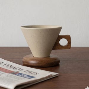 Könnte beinhalten: Ein beigefarbener Keramik-Kaffee-Pour-Over mit Holzgriff und -sockel. Der kegelförmige Filterhalter sitzt auf einem runden Holzsockel. Der Griff hat eine kreisförmige Öffnung. Eine Zeitung ist im Vordergrund.