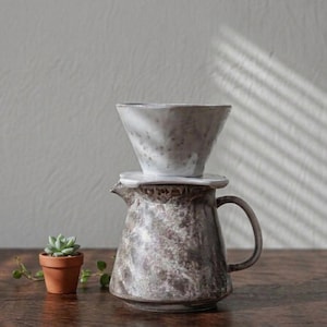 May include: A set of three ceramic mugs with a speckled, matte finish. The mugs are in shades of cream, light gray, and dark gray. Each mug has a simple, minimalist design and a rounded handle.