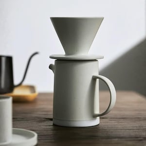 Puede incluir: Un juego de cafetera de cerámica para verter de color crema. Incluye un vertedor en forma de cono, una base circular y una jarra cilíndrica con asa. El diseño es minimalista y moderno, adecuado para preparar café en casa.
