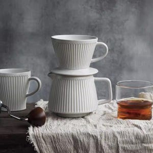 May include: A white ceramic coffee set with vertical ribbed detailing. The set includes a coffee pot, a filter holder, and two matching mugs. A glass of amber liquid sits nearby, suggesting the set is for brewing and enjoying coffee.