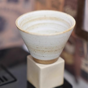 May include: A handmade, off-white ceramic bowl with a conical shape, resting on a small, square, cream-coloured stand. The bowl has a speckled texture and a light brown rim and base. The interior shows concentric circles.