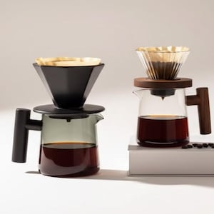 Puede incluir: Dos juegos de café pour-over. Uno tiene un portafiltro negro angular y una jarra de vidrio teñido de gris con un asa negra. El otro tiene un portafiltro de vidrio transparente con una base de madera y una jarra de vidrio transparente con un asa de madera.