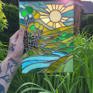 May include: Stained glass panel showing a landscape with a bright yellow sun, green rolling hills, and a blue river. The artwork features trees with green circular leaves and a clear sky. The panel is held up in front of a background of green grass.
