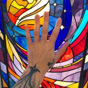 Op de afbeelding: Een kleurrijk glas-in-loodraam met een levendig ontwerp. Het kunstwerk toont een cirkelvormig zonachtig element in geel, oranje en rood, omgeven door blauwe en paarse secties. Een hand bevindt zich voor het glas.