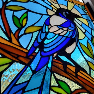 May include: Stained glass panel featuring a blue and white magpie perched on a brown branch. The bird is set against a backdrop of blue, yellow, and green glass, creating a vibrant and colorful artwork. The panel is framed in black.