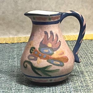 May include: A small ceramic pitcher with a light pink base color, featuring a hand-painted stylized bird motif in shades of blue, yellow, and green. The pitcher has a curved handle with blue stripes and a blue rim around the top and base.