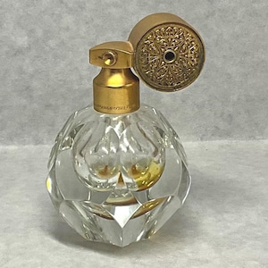 May include: A clear glass perfume bottle with a faceted, diamond-like base and a gold-toned neck. The bottle features a round, gold-toned cap with an intricate, embossed floral pattern on its top surface, reflecting a classic and ornate design.
