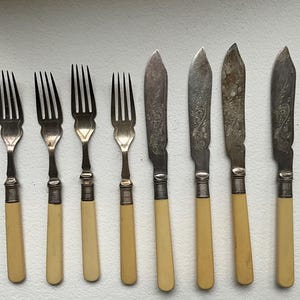 May include: A collection of antique silverware, including forks and butter knives. The forks have three tines and cream-colored handles. The knives have ornate designs on the blades and matching handles. The silverware is arranged on a white surface.