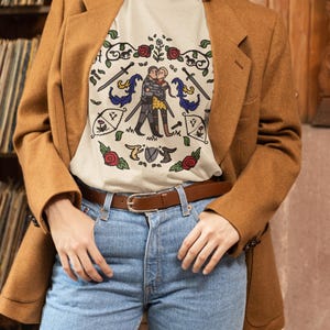 May include: A cream-colored t-shirt featuring a central illustration of two figures embracing, surrounded by a decorative border of red roses, swords, small horses, and banners. The design is rendered in a line-art style with muted colors.